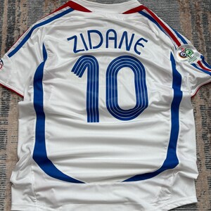 May include: White soccer jersey with blue and red accents. The jersey features the name "ZIDANE" and the number "10" in blue on the back. A logo is visible on the right sleeve. The jersey has a v-neck and blue stripes.