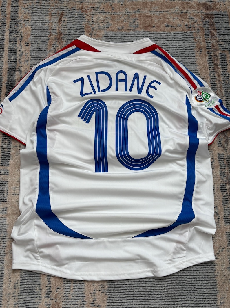 May include: White soccer jersey with blue and red accents. The jersey features the name "ZIDANE" and the number "10" in blue on the back. A logo is visible on the right sleeve. The jersey has a v-neck and blue stripes.