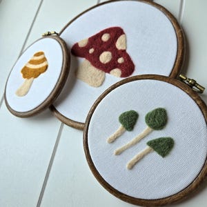May include: Three circular wooden embroidery hoops containing white fabric with needle-felted mushroom designs. One hoop features a large red mushroom with cream spots, another a yellow-striped mushroom, and the third shows three small green mushrooms.