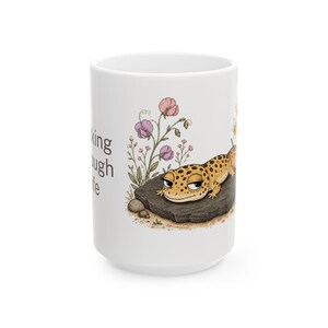 Może przedstawiać: Biały ceramiczny kubek z ilustracją gekona z kreskówki. Żółto-brązowy gekon leży na skale z kwiatami. Tekst na boku kubka brzmi "king tough life".