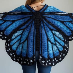 Op de afbeelding: Een gehaakte sjaal ontworpen als vlindervleugels, met een levendig verloop van blauwtinten van licht naar donker, omlijnd met dik zwart garen en geaccentueerd met kleine witte stippen langs de geschulpte randen.