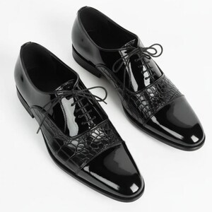 May include: A pair of black dress shoes featuring a high-gloss patent finish, a lace-up closure with thin black laces, and a textured crocodile-embossed leather panel across the vamp and toe cap, set against a plain white background.
