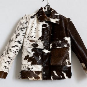 Peut inclure: Veste patchwork avec un motif de peau de vache. La veste présente un mélange de sections tachetées blanches et noires avec des sections marron et noires. Elle a un col, des boutons et deux poches avant. La veste est suspendue à un cintre noir.