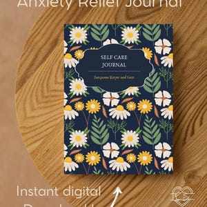 Könnte beinhalten: Ein floral gemustertes Journal mit dem Text "Anxiety Relief Journal" und "Self Care Journal". Das Cover zeigt weiße und gelbe Gänseblümchen und grüne Blätter auf marineblauem Hintergrund. Das Journal liegt auf einer Holzoberfläche.