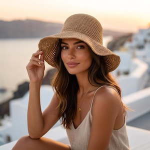 Peut inclure: Un chapeau de soleil en paille tressée avec un large bord. Le chapeau est de couleur beige clair et a une couronne arrondie. Le chapeau est tenu par une personne. L'arrière-plan présente un paysage côtier.