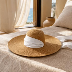 Peut inclure: Un chapeau de soleil &agrave; larges bords avec une couronne et un bord tiss&eacute;s bruns, et un bandeau en tissu blanc. Le chapeau repose sur une surface beige, avec une vue sur l'oc&eacute;an en arri&egrave;re-plan. Le chapeau est con&ccedil;u pour la protection solaire.