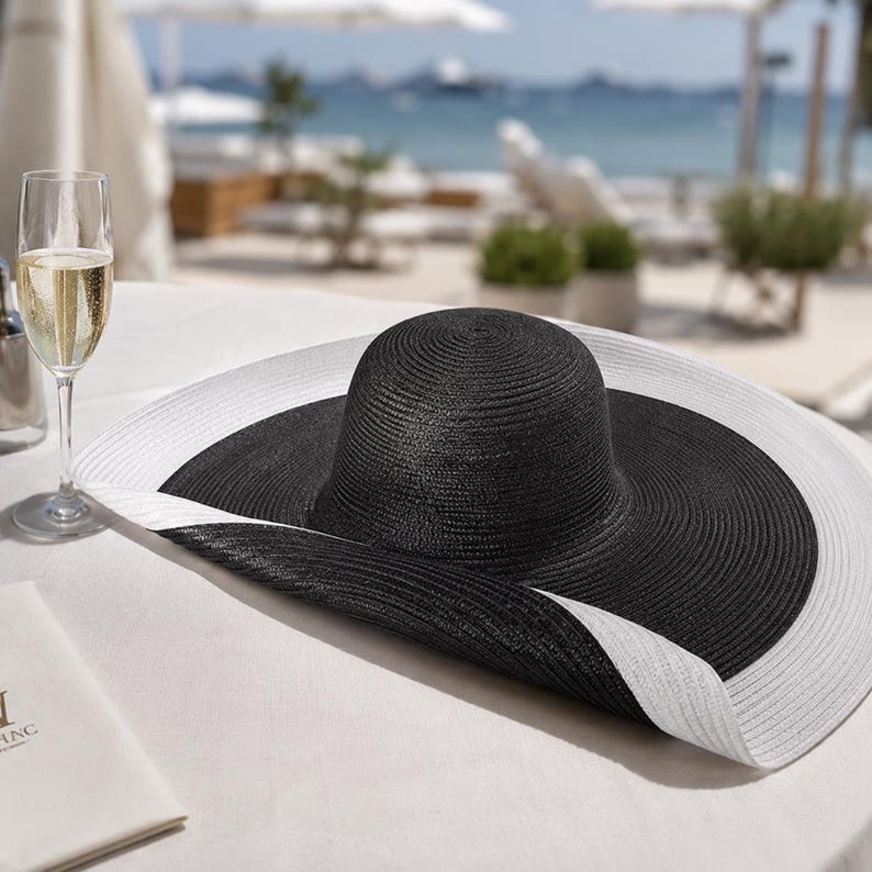 May include: A wide-brimmed sun hat with a black crown and a white brim rests on a white tablecloth. A glass of champagne sits nearby. The hat is a stylish accessory for sunny days at the beach or a resort.