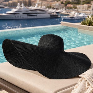 May include: A wide-brimmed black sun hat rests on a beige lounge chair near a pool. The hat has a rounded crown and a broad, gently curved brim. The background features a body of water with boats and buildings.
