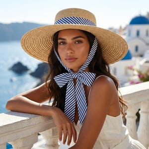 May include: A straw hat with a navy and white striped ribbon and ties. The hat has a round crown and a wide brim. The ties are tied in a bow under the chin. The background shows a coastal scene with blue water and white buildings.
