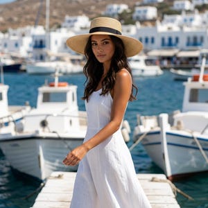 Peut inclure: Une femme porte une robe d'été blanche et un chapeau de paille avec un ruban noir, debout sur un ponton en bois. En arrière-plan, des bateaux et des bâtiments près de l'eau.