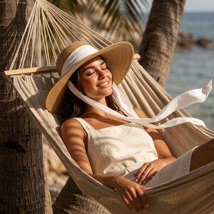 Peut inclure: Une femme portant un chapeau de paille &agrave; larges bords avec un ruban blanc, se d&eacute;tendant dans un hamac beige. Elle porte une robe cr&egrave;me. L'arri&egrave;re-plan comprend des palmiers et l'oc&eacute;an.