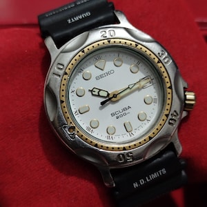 Puede incluir: Un reloj Seiko Scuba 200m de cuarzo plateado y dorado con esfera blanca, correa de caucho negra y detalles dorados. La esfera muestra el logotipo de Seiko, números romanos y el texto "SCUBA 200m". La correa está marcada con "N.D. LIMITS".