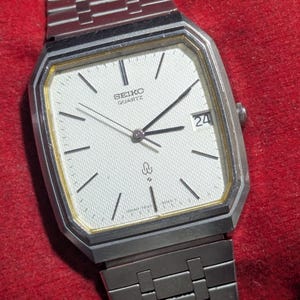 Op de afbeelding: Een zilverkleurig Seiko Quartz polshorloge met een rechthoekige kast, een witte wijzerplaat met textuur en een metalen schakelarmband. Het horloge heeft zwarte uurmarkeringen, dunne zwarte wijzers, een datumvenster met het getal 24 en een goudkleurige rand.
