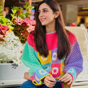 May include: A long-sleeved crochet sweater featuring a vibrant patchwork design of square motifs in bright pink, yellow, light blue, green, and purple. Each square contains a white flower pattern. The sweater has a crew neckline and loose sleeves.