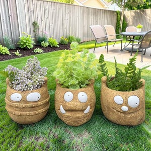 May include: Three whimsical, tan-colored monster-faced planters filled with greenery, set on a green lawn. Each planter has a unique expression with white eyes and a mouth. The planters are of varying heights and shapes.