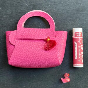 May include: A bright pink miniature handbag with a handle and a red tassel. Next to it is a tube of strawberry lip balm and a small pink duck figurine. The lip balm tube reads "Strawberry Lip Balm".