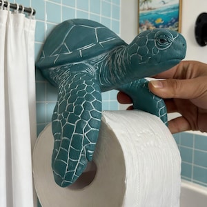 May include: A teal sea turtle wall-mounted toilet paper holder with a textured shell and flipper design. The turtle is sculpted with detailed scales and holds a standard roll of white toilet paper between its front flippers, extending outward.