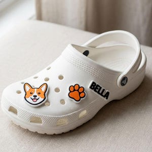 May include: A white foam clog shoe featuring three decorative charms: a colorful Corgi dog face, an orange paw print, and a black rectangular charm with the name BELLA printed in white capital letters on the side of the shoe's upper surface.