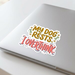 May include: A die-cut sticker featuring the text MY DOG RESTS I OVERTHINK in brown and coral pink lettering. The design includes small illustrations of dog bones and paw prints in light green and brown, all set against a white background.