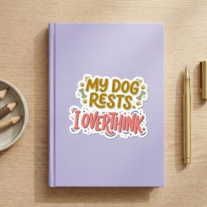 May include: A light purple notebook featuring a central sticker with the text "MY DOG RESTS. I OVERTHINK" in brown and pink, accented by small green bone and paw print illustrations, accompanied by a gold pen and three pencils in a small bowl.