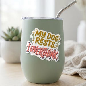 May include: A sage green stainless steel tumbler with a clear lid and a silver metal straw. A sticker on the front features the text "MY DOG RESTS I OVERTHINK" in yellow and pink, surrounded by small decorative paw prints and bone icons.