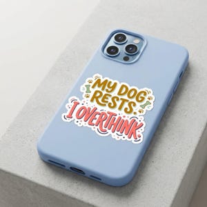 May include: A light blue smartphone case featuring a central sticker with the text "MY DOG RESTS I OVERTHINK" in brown and pink lettering, accented by small green bone icons and brown paw prints, with a triple-lens camera cutout at the top.