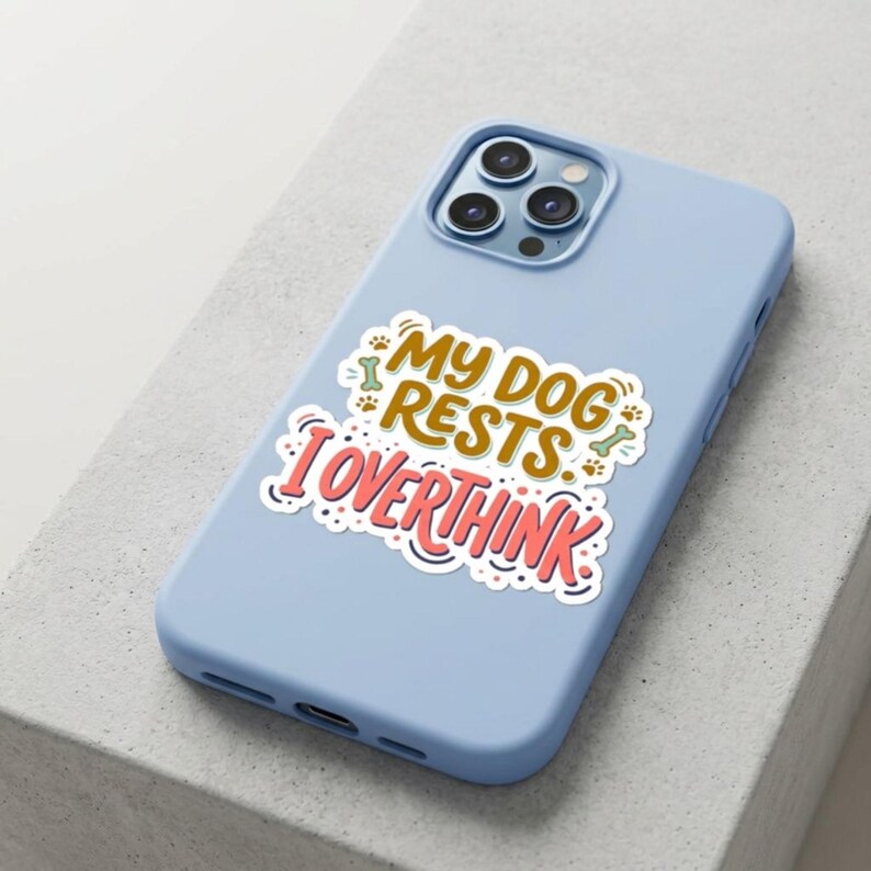 May include: A light blue smartphone case featuring a central sticker with the text "MY DOG RESTS I OVERTHINK" in brown and pink lettering, accented by small green bone icons and brown paw prints, with a triple-lens camera cutout at the top.