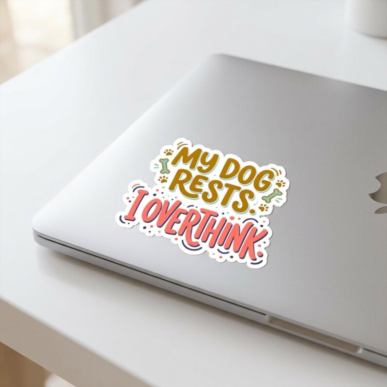 May include: A die-cut sticker featuring the text MY DOG RESTS I OVERTHINK in brown and coral pink lettering. The design includes small illustrations of dog bones and paw prints in light green and brown, all set against a white background.