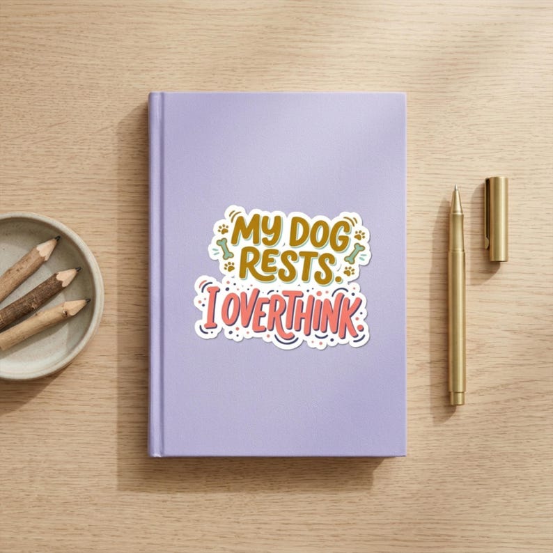May include: A light purple notebook featuring a central sticker with the text "MY DOG RESTS. I OVERTHINK" in brown and pink, accented by small green bone and paw print illustrations, accompanied by a gold pen and three pencils in a small bowl.