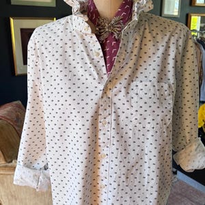 Può includere: Camicia bianca con un piccolo motivo floreale scuro, colletto a balze e taschino sul petto. Visibili una sciarpa bordeaux e una spilla decorativa. Il manichino è etichettato "MODEL 1995".
