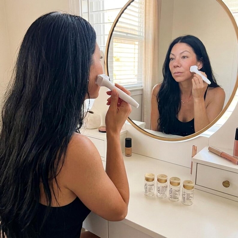 Puede incluir: Una mujer usa un rodillo facial blanco en su mejilla frente a un espejo. El espejo es redondo con un marco dorado. Varias botellas de vidrio peque&ntilde;as con tapas doradas est&aacute;n en el tocador. La mujer tiene el pelo largo y negro y lleva una camiseta sin mangas negra.