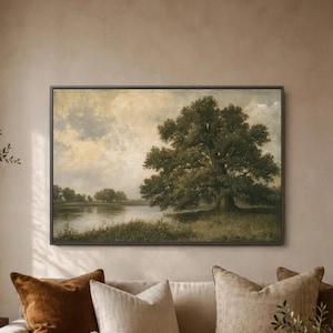 May include: A rectangular landscape painting in a dark frame depicting a large, leafy tree standing on the grassy bank of a calm river. The sky is filled with soft, muted clouds in shades of beige and cream, casting a gentle light over the scene.
