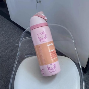 May include: A light pink Owala water bottle featuring Hello Kitty graphics and a peach-colored product label. The label includes the text: Hi there. I'm leak proof. I'm insulated. And I have a built-in straw. The bottle has a pink lid and handle.