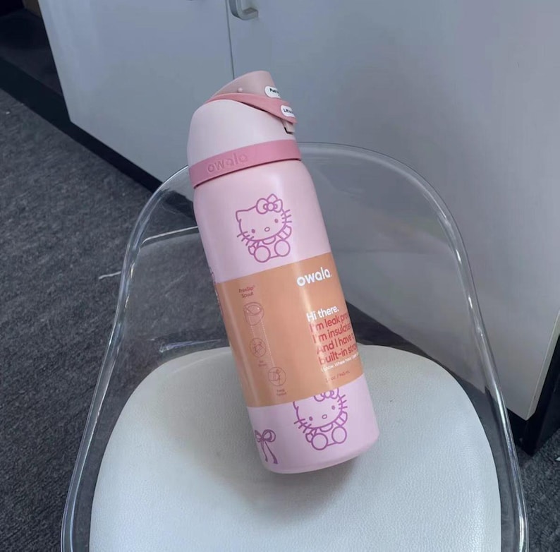 May include: A light pink Owala water bottle featuring Hello Kitty graphics and a peach-colored product label. The label includes the text: Hi there. I'm leak proof. I'm insulated. And I have a built-in straw. The bottle has a pink lid and handle.