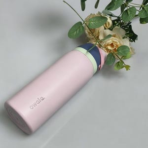 May include: A light pink insulated water bottle with a multi-colored lid, featuring a navy blue, light green, and pink design. The bottle has the word "owala" printed on the side. The bottle is next to a bouquet of flowers.