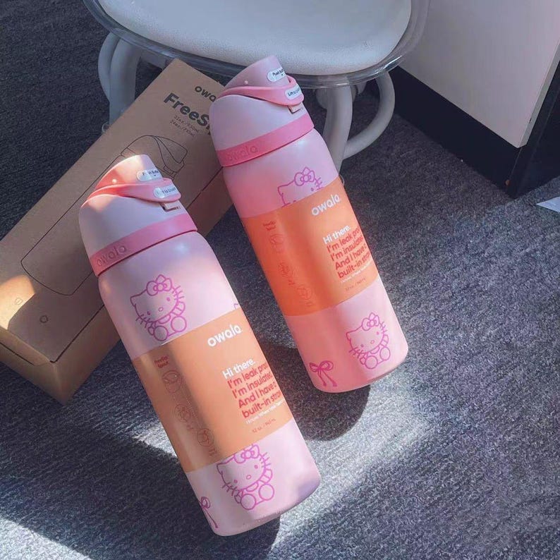 May include: Two light pink Owala FreeSip water bottles featuring Hello Kitty graphics and a peach-colored band with the text: Hi there, I'm leak proof, I'm insulated, And I have a built-in straw. Each bottle has a pink lid with a push-button mechanism.