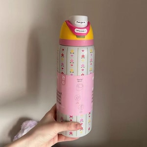 May include: A floral-patterned water bottle with a yellow and pink lid. The bottle has a pink band with text and illustrations. The lid has the text "Push But" and "Lift to Unle". The bottle is designed to keep drinks cold for 24 hours.