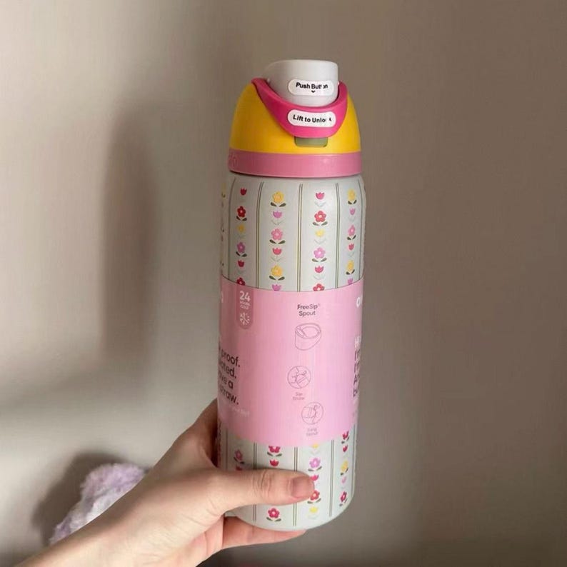 May include: A floral-patterned water bottle with a yellow and pink lid. The bottle has a pink band with text and illustrations. The lid has the text "Push But" and "Lift to Unle". The bottle is designed to keep drinks cold for 24 hours.