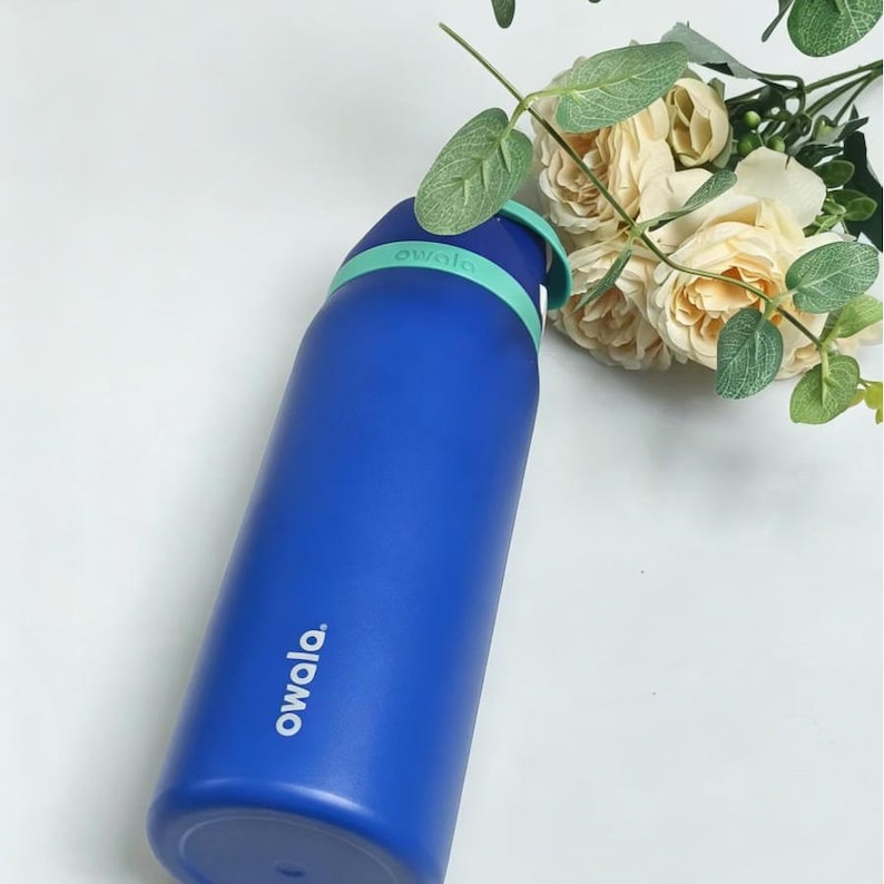 May include: A blue Owala water bottle with a mint green handle and lid. The bottle has the word "owala" printed on the side in white. The background includes white flowers and green leaves.