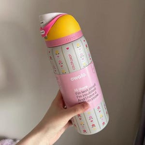 May include: A pink and white insulated water bottle with a yellow top. The bottle has a floral pattern and the text "COLOR DROP" and "owala". The bottle also has the text "Hi there. I'm leak proof. I'm insulated. And I have a -in straw."