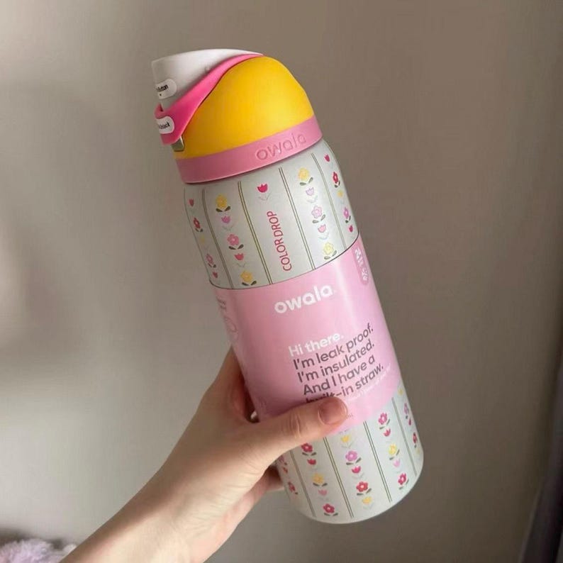 May include: A pink and white insulated water bottle with a yellow top. The bottle has a floral pattern and the text "COLOR DROP" and "owala". The bottle also has the text "Hi there. I'm leak proof. I'm insulated. And I have a -in straw."