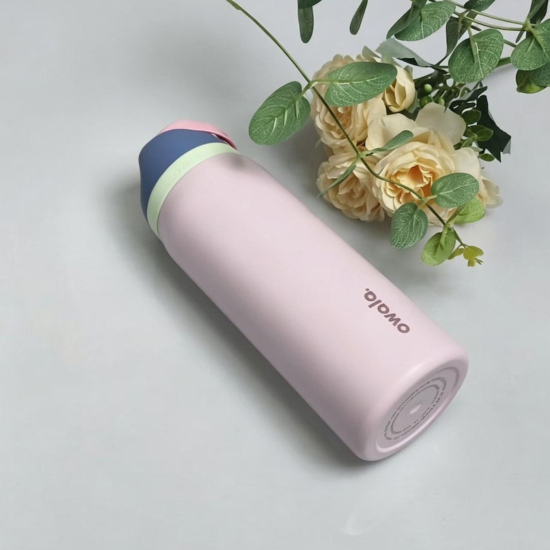 May include: A light pink insulated water bottle with a blue and pink lid. The bottle has the word "owala" printed on the side. The bottle is cylindrical and appears to be made of metal. The background includes flowers and greenery.