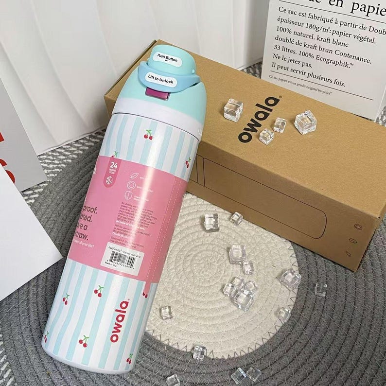 May include: A stainless steel Owala water bottle with a light blue and white striped pattern, adorned with cherry illustrations. The bottle features a pink band with product information and a blue lid with a push button and "Lift to Unlock" text.