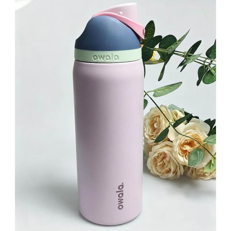 May include: A light pink Owala water bottle with a blue and green lid and a pink handle. The bottle has the word "owala" printed on it in white. The bottle is next to a bouquet of cream-colored roses.