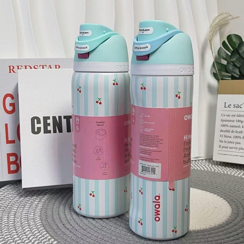 May include: Two Owala water bottles. The bottles have a light blue and white striped pattern with small cherry illustrations. The lids are light blue with a push button and a lift-to-unlock feature. Pink labels wrap around the bottles.