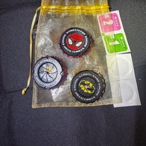 May include: Three circular engine start-stop button covers featuring graphic designs of a red spider web, a black and yellow bat symbol, and a black and white skull. The covers are displayed with a yellow organza drawstring bag and cleaning wipes.
