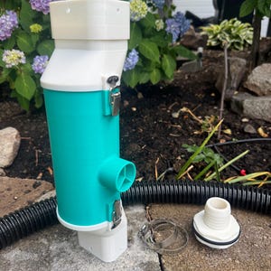 May include: A teal and white cylindrical debris filter housing with metal latches, featuring a side port and top and bottom square-shaped connectors. Included are a black corrugated hose, two metal hose clamps, and a white threaded adapter.