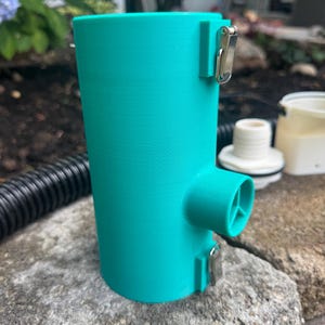 May include: A cylindrical teal plastic housing component featuring two metal toggle latches on the side and a protruding circular port with an internal three-spoke support structure. The object has a smooth, matte finish and a vertical seam.