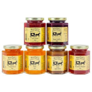 May include: A collection of six glass jars of Rachel's Texas Kitchen jam and apple butter. The jars have gold lids and labels with a stagecoach logo. Flavors include Texas Twister, Texas Traffic, Tropical Storm, Peach, Dutch Apple Butter, and Strawberry Rhubarb.