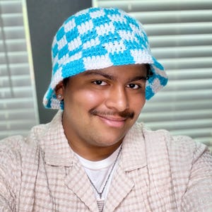 May include: A blue and white checkered crochet bucket hat. The hat features a classic checkerboard pattern in light blue and white. The hat is handmade and has a brim.